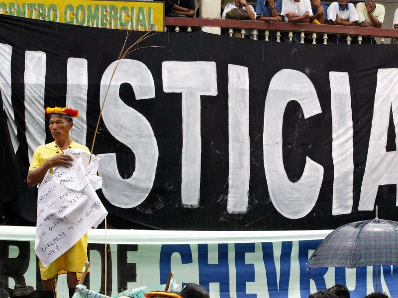 A Secoya elder stands in front of a banner reading 'Justice' during a demonstration at the start of the trial against Chevron in Lago Agrio, Ecuador. Photo Credit: Lou Dematteis, from Crude Reflections